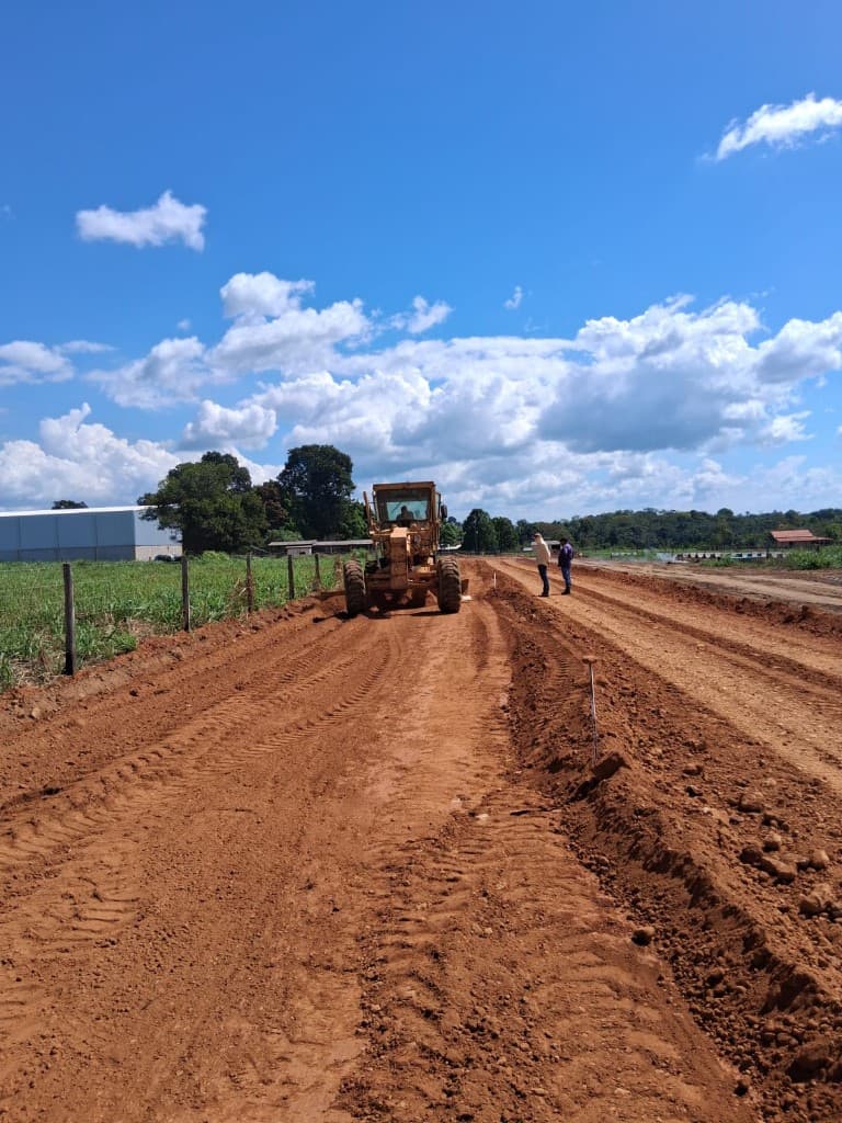 Motoniveladora amarela nivelando solo avermelhado com equipe técnica acompanhando a obra